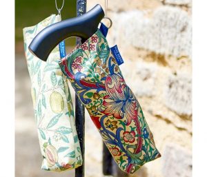 Image is a photograph of a folded walking stick inside a William Morris-inspired print, hanging against a sandstone wall