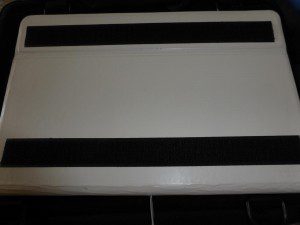 Photograph shows the back of a white tablet computer case, with two parallel strips of black adhesive hook tape on the back.