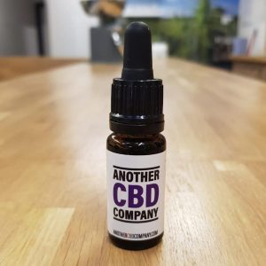 Image is a photograph of a tincture bottle of Indica CBD oil on a wooden table